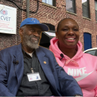 An older gentleman in a black shirt, blue blazer, and Air Force hat sits next to a woman in a pink Nike hoodie. The background is an image of MCVET's building.